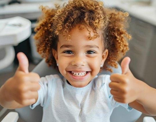 Child in dental chair, giving two thumbs-up