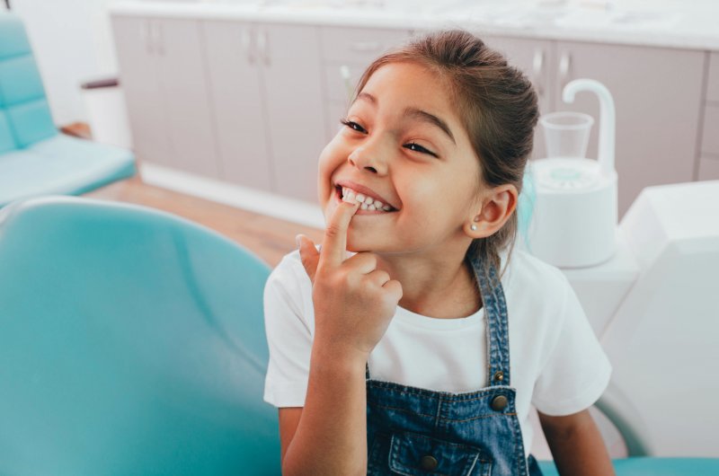 a little girl smiling and pointing to her teeth while in the dentist’s chair